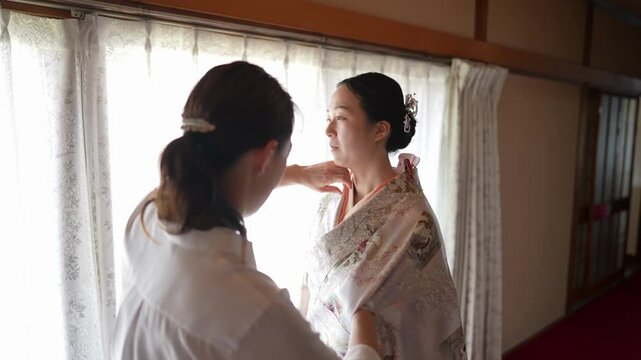 An assistant adjusts the overlapping collar of a woman's patterned kimono in soft window light. Japan, April 19, 2025, spring.