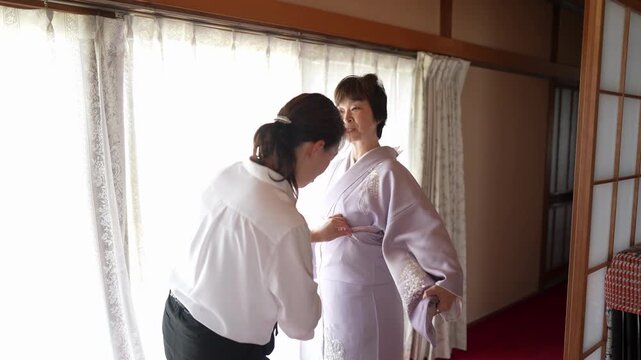 An assistant bends forward to secure a pink tie around a woman's lavender kimono. Japan, April 19, 2025, spring.