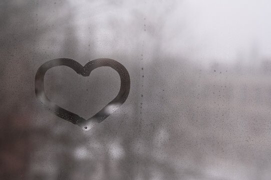 Heart shape drawn on wet gray glass window pane covered with condensation water drops. Romantic heart symbol looks positive on dark moody rainy backdrop. Melancholy romance concept.