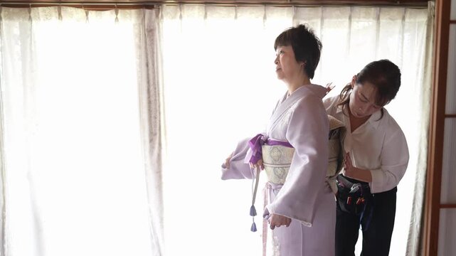 A woman stands in profile while an assistant adjusts the obi at the back of her lavender kimono. Japan, April 19, 2025, spring.