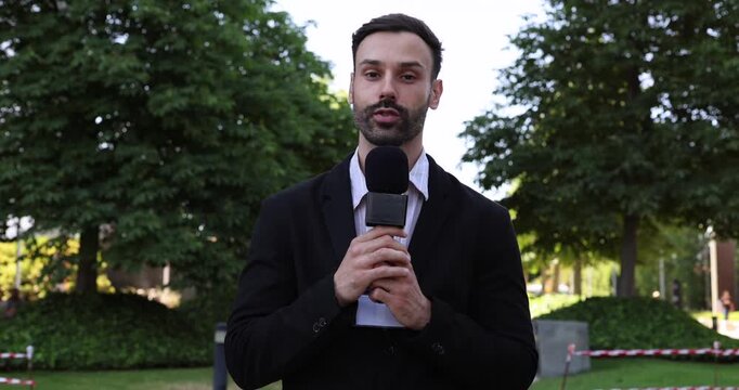 Male journalist in a suit speaking into a microphone while reporting live from an outdoor location, with green trees in the background creating a professional and serious atmosphere