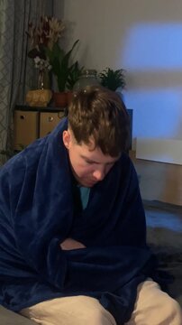 Adult ill guy talking paper handkerchief and sneezing while sitting on sofa covered with blanket.