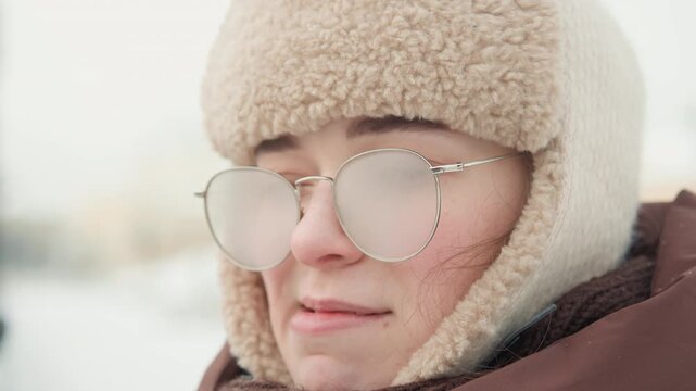 Close-up eye-level shot of young woman wearing shearling-lined knit hat, round metal glasses with frosted lenses, quilted brown jacket and scarf, breathing visible cold air and shifting from pursed