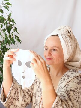 Woman with towel on head holding facial sheet mask in front of her, smiling, preparing for home spa skincare routine