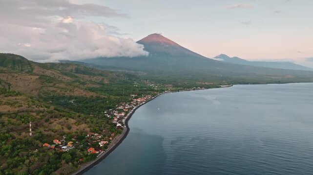 Mount Agung rising above low cloud band at morning sunrise overlooking Amed beach shoreline and its coastal village. Calm sea and tranquil tropical panorama of Bali island. Aerial view drone flight