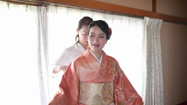 A smiling woman extends her orange kimono sleeves and looks back toward the assistant at her waist. Japan, April 19, 2025, spring.