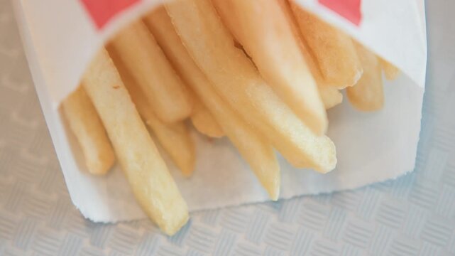 Top-down close-up showing pale golden French fries spilling from white paper container while blurred fingertip reaches in to pick up fry; soft diffused overhead light creates gentle highlights