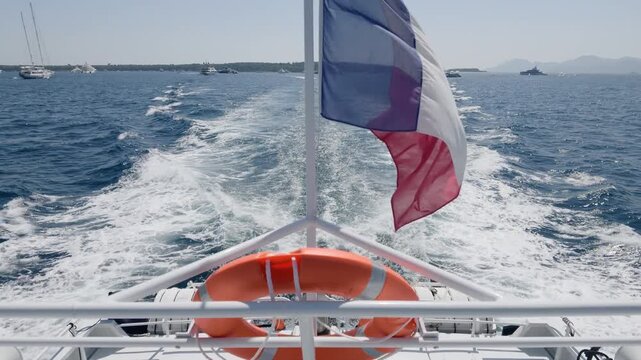 Close-up of a French flag flying at a yacht's stern with wake and yachts near a wooded coastline, Ile Sainte-Marguerite, France