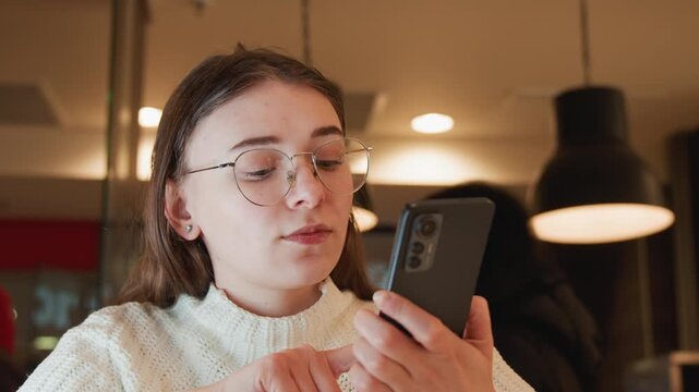 Young woman in metal rim glasses sits inside cozy cafe, focused on smartphone before pausing to rest head on hand with thoughtful expression, soft warm light and calm lifestyle mood