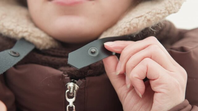 Close-up view shows lower face, hands, and upper chest as person fastens snaps and zipper on brown quilted winter jacket with beige faux fur hood trim, with straps hanging and fabric folding