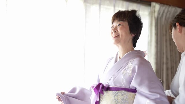A smiling woman in a lavender kimono receives help adjusting her obi at the back. Japan, April 19, 2025, spring.