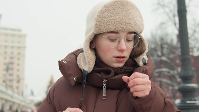 Smiling young woman in beige parka, knit trapper hat, and clear glasses lowers face mask with both hands in winter park, showing relief and friendliness in soft daylight near bridge and snowy trees
