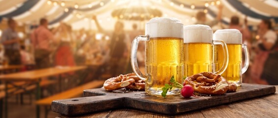 Drei Maßkrüge mit frischem Bier und Brezeln auf einem rustikalen Holzbrett. Unscharfer Hintergrund mit feiernden Menschen in einem Oktoberfest-Bierzelt. Breites Panorama-Banner mit Freiraum