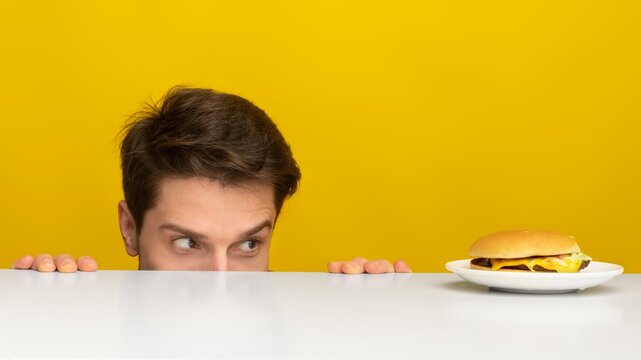 Curious Young Man With Light Skin Looking At A Tasty Hamburger On Table, In Studio Setting, Evoking Hunger And Anticipation For Delicious Food. Fast Food And Healthy Eating Concept.