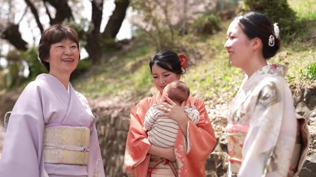 A smiling woman in an orange kimono holds a baby facing her beside women in lavender and patterned kimonos. Japan, April 19, 2025, spring.