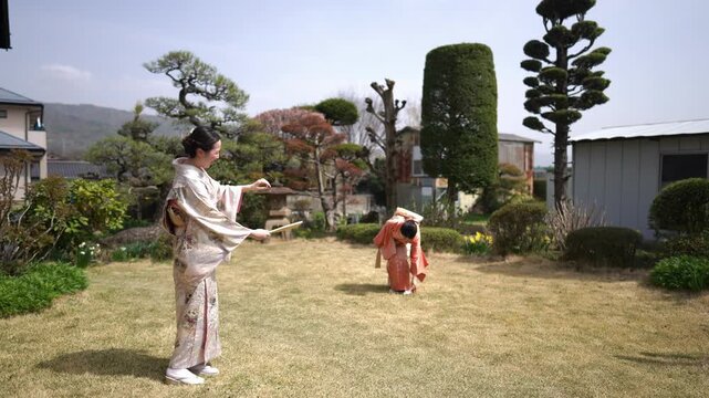 Two women in patterned and orange kimonos face one another holding wooden paddles on a traditional garden lawn. Japan, April 19, 2025, spring.