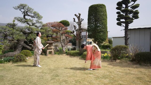 A woman in an orange kimono bends her knees with a wooden paddle as another kimono-clad woman waits across the lawn. Japan, April 19, 2025, spring.