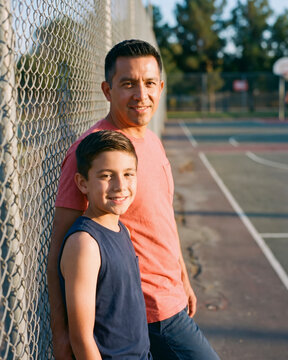 Resolute father and son standing by court fence on sunny basketball court