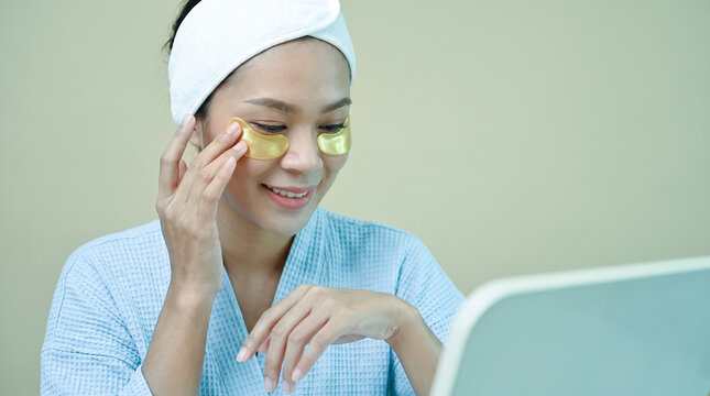 Smiling Young Asian Woman in Bathrobe Applying Gold Under-Eye Patches
