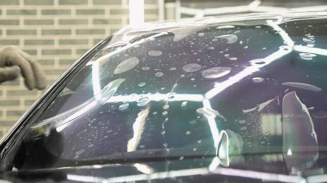 Hands of a man glue a clear protective film against scratches on a car windshield close up. This is an athermal window tint wrap. Worker installs a chameleon vinyl sheet on an automobile glass. 