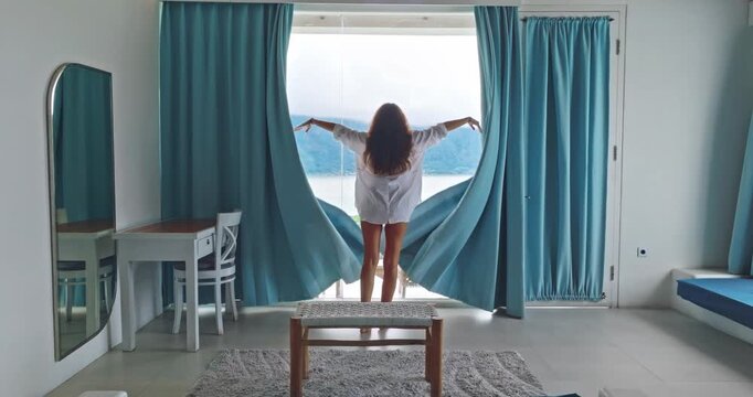 Woman opening teal curtains in a bright hotel room, letting sunlight flood the space, symbolizing new beginnings, travel, and vacation relaxation during a peaceful morning. Batur lake, Bali, Indonesia