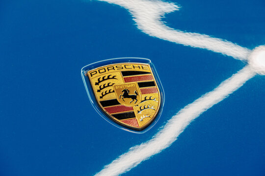 Golden luxury Porsche logo badge on a blue metallic hood bonnet. Premium German automobile corporate emblem suitable for sport car models like Carrera, GT3, Cayman, Boxster, Taycan, Panamera, Macan.