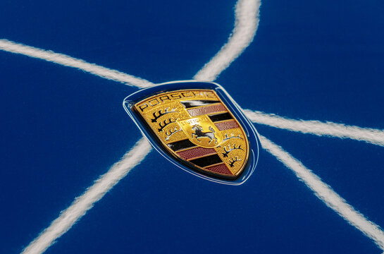 Golden luxury Porsche logo badge on a blue metallic hood bonnet. Premium German automobile corporate emblem suitable for sport car models like Carrera, GT3, Cayman, Boxster, Taycan, Panamera, Macan.