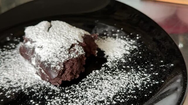 A teaspoon takes a bite of soft chocolate brownie dusted with powdered sugar on a black plate, capturing an appetizing moment of enjoying a homemade dessert. Close up.