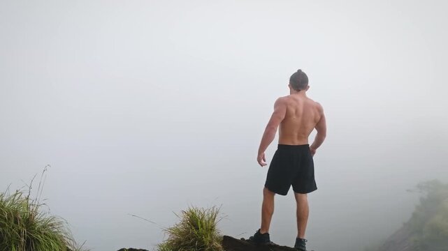 Muscular man with braided hair standing shirtless on a mountain peak, looking at the expansive foggy landscape, embodying a sense of achievement and strength after an adventurous hike