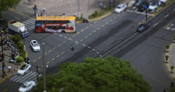 Cars and yellow taxi drive through a busy city crossroads with green trees in daytime, recorded in tilt shift miniature timelapse