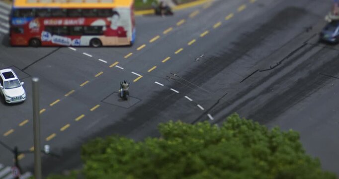 Vehicles and cars move through an urban street crossroads beside green trees, recorded in miniature tilt shift timelapse