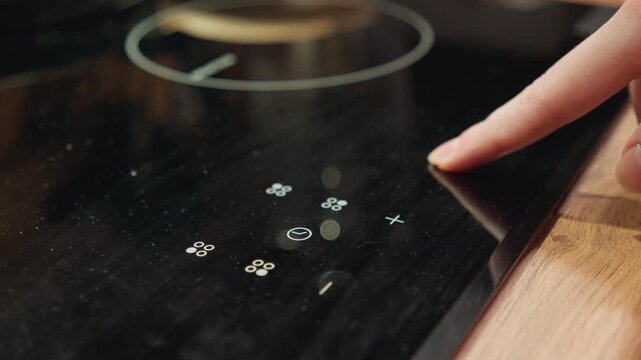 Close view shows index finger pressing glowing touch control on glossy black induction cooktop, with circular heating zone, reflective glass surface, and wooden countertop creating clean modern