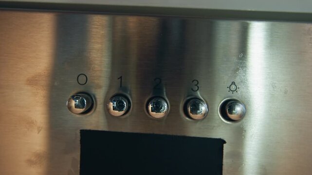 Eye-level view of brushed stainless steel appliance control panel with five round chrome buttons above dark rectangular display as fingertip presses one control, showing reflective surface and