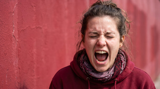 Young woman screaming with eyes closed against a red wall for intense anger and emotional stress concept