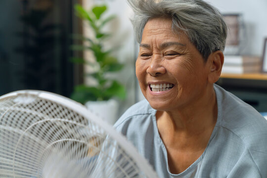 Cheerful elderly Asian woman sitting on a sofa and turning on a fan to cool down during extremely hot weather at home.