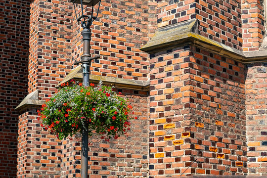 Historic red brick gothic church wall featuring stone ledge, with a black metal street lantern post decorated with a hanging pot of blooming red and white flowers.