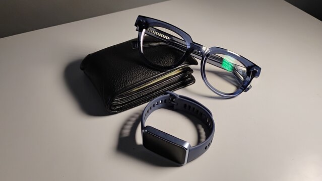 A black leather wallet, dark blue eyeglasses, and a dark blue fitness tracker watch arranged on a clean white table.