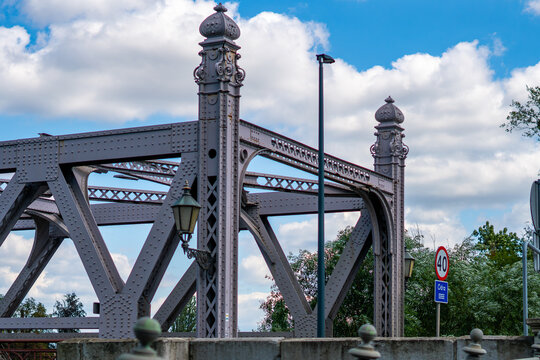 Historic Piast Bridge in Brzeg featuring riveted steel truss construction with ornate decorative turrets, lanterns, street light pole, and speed limit sign under a blue cloudy sky.