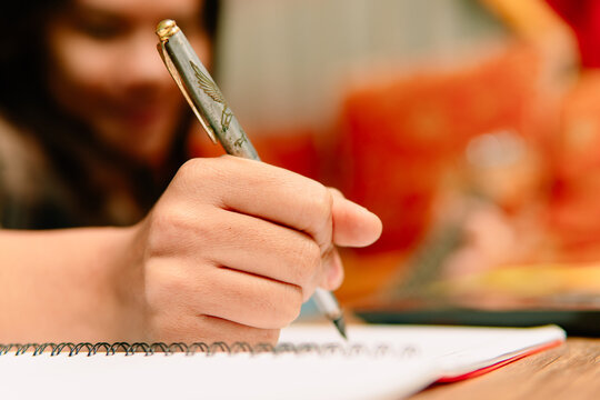 Macro close-up of Asian woman hand holding pen writing on spiral notebook page indoors.
