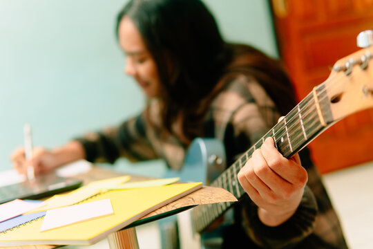 Close-up of an Asian woman hand holding electric guitar neck while writing lyrics at a desk.
