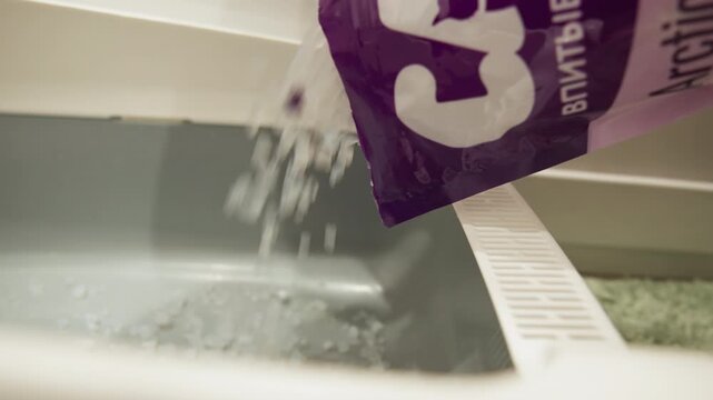 Close-up low-angle view of powdered and sheet detergent being dispensed into open washing machine drum, visible cascade of powder, torn sheet fragments and floating particles creating subtle ripples