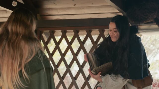 Two women stand face-to-face inside rustic wooden shelter while woman on right raises torn paper and holds open leather-bound notebook, speaking with animated gesture, forward lean, and engaged