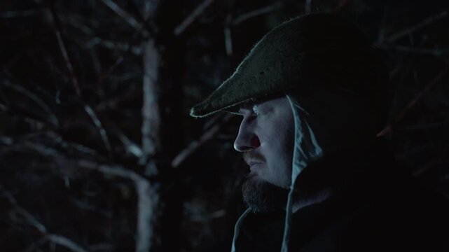 Close side portrait of man wearing earflap hat standing in snowy forest at night, visible breath, downward gaze, cool blue light, warm red glow, and quiet contemplative winter atmosphere