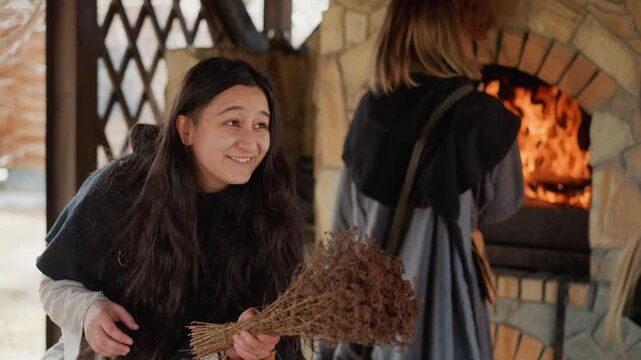 Foreground woman leans forward smiling, holding rustic twig broom while background woman reaches into stone wood-fired oven with orange flames, showing active stoking and sweeping motion in cozy