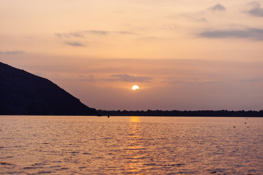 The sun rises through soft clouds over Lake Skadar, casting a warm golden reflection across the calm water beside a dark silhouetted hillside. Montenegro nature
