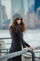 Woman looking over shoulder near waterfront fence in New York. An adult female in dark clothing and knit cap stands by a river embankment. © Vadim
