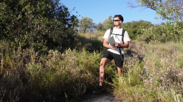 Adventurous young man with a backpack walking on a trail, stopping to check his route and orient himself using a paper map in a sunny forest