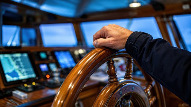 A hand gripping a wooden ship steering wheel inside a vessel&rsquo;s cockpit with digital navigation screens in the background.
