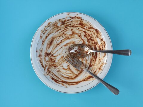 Dirty white ceramic plate with peanut sauce leftover, spoon, and fork isolated on cyan blue background. Concept of finished meal, food waste, dirty dishes, or delicious dining experience