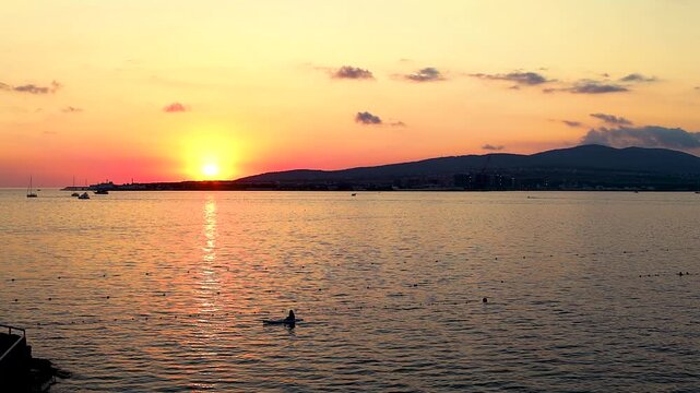 Sunset over calm water with boats sailing, silhouetted trees framing the scene, and distant hills creating a serene landscape in a coastal region